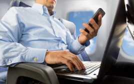 אדם משתמש בטלפון ומחשב נייד במטוס. אילוסטרציה (צילום: אינגאימג')