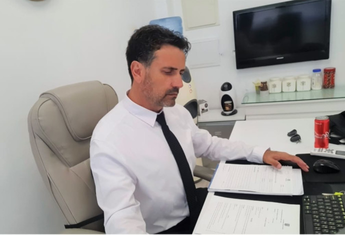 A man in a white shirt and black tie sits at a desk, reviewing documents. A computer and a can of soda are nearby.